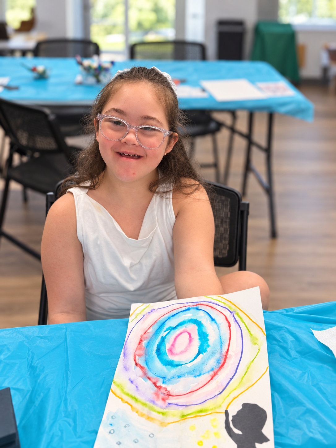 A smiling girl at a craft table holding up a colorful watercolor painting she made